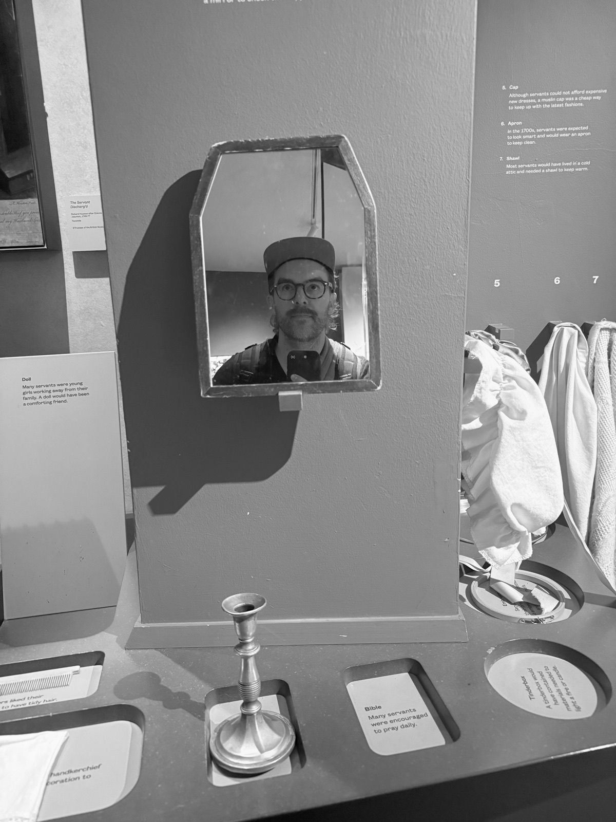 Colin Hanner, wearing a cap and glasses, reflected in a small angular mirror mounted on a museum exhibit wall, with display labels and objects around it.